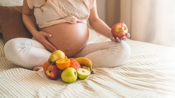Eine schwangere Frau sitzt auf einem Bett, vor ihr ein Teller mit Obst
